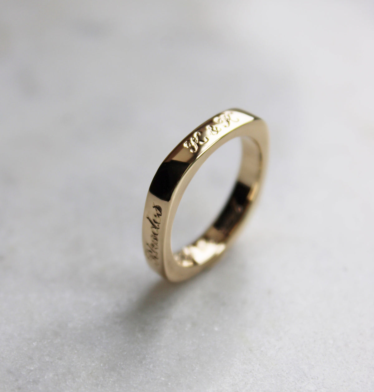 A gold band ring with engraved text, standing upright on a light gray textured surface.